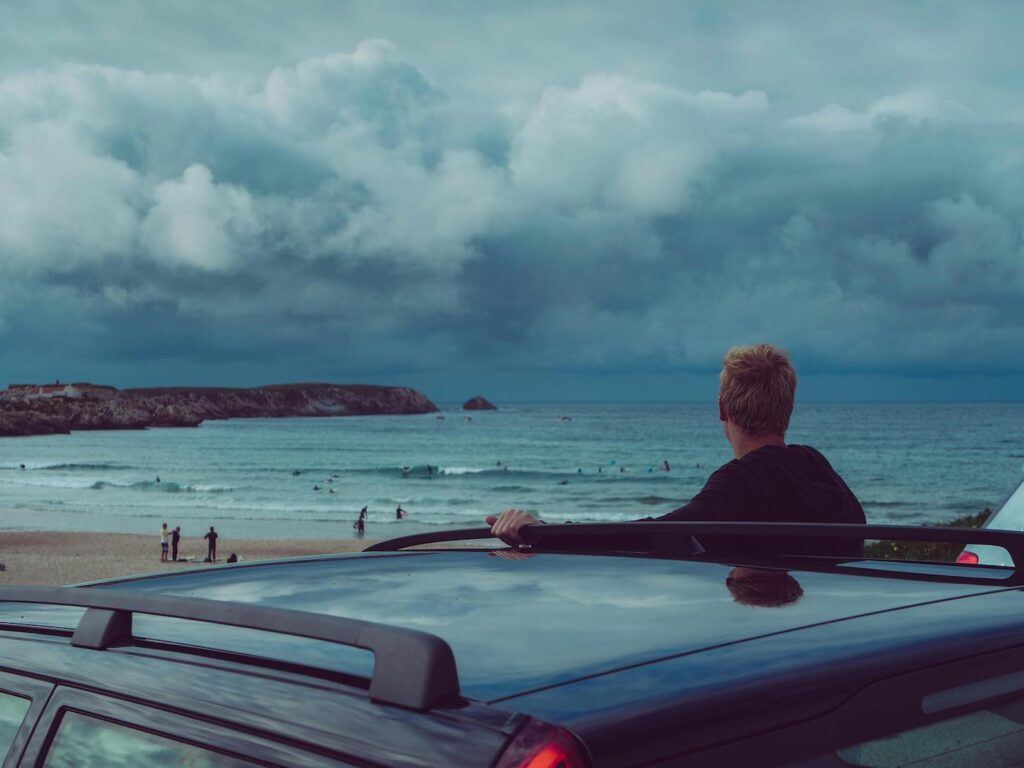 A person stands beside a car looking at surfers entering teh water on a beach with a cloudy sky