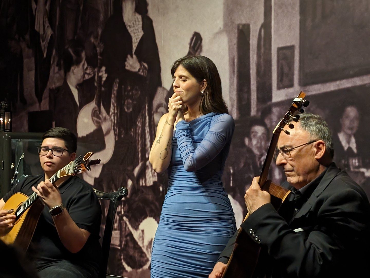 A female Fado singer clasps he hands with her eyes closed, as two guitar players accompany her