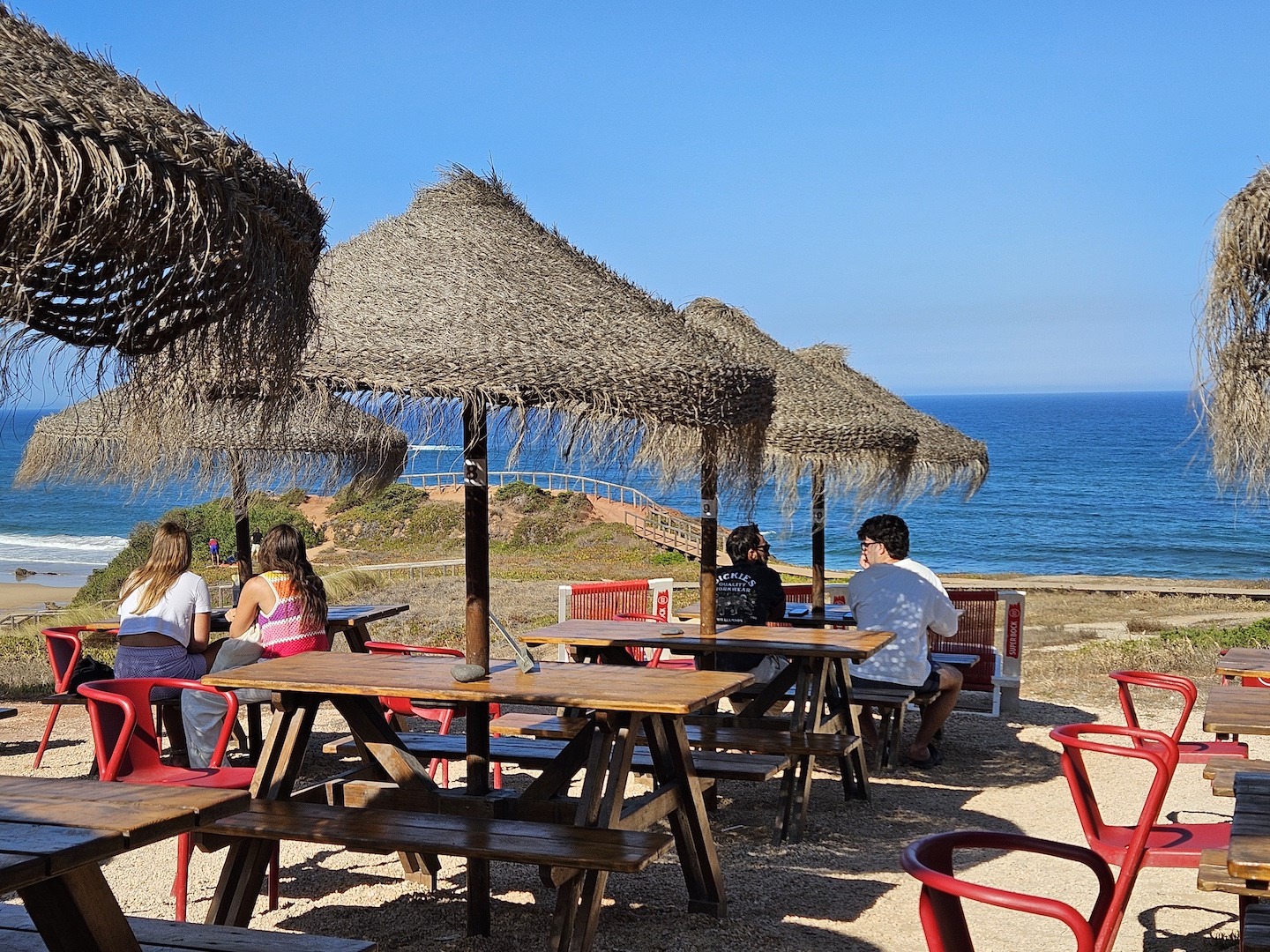 Four people sit at two different tables at a small outdoor cafe overlooking Amadora beach in Sagres.