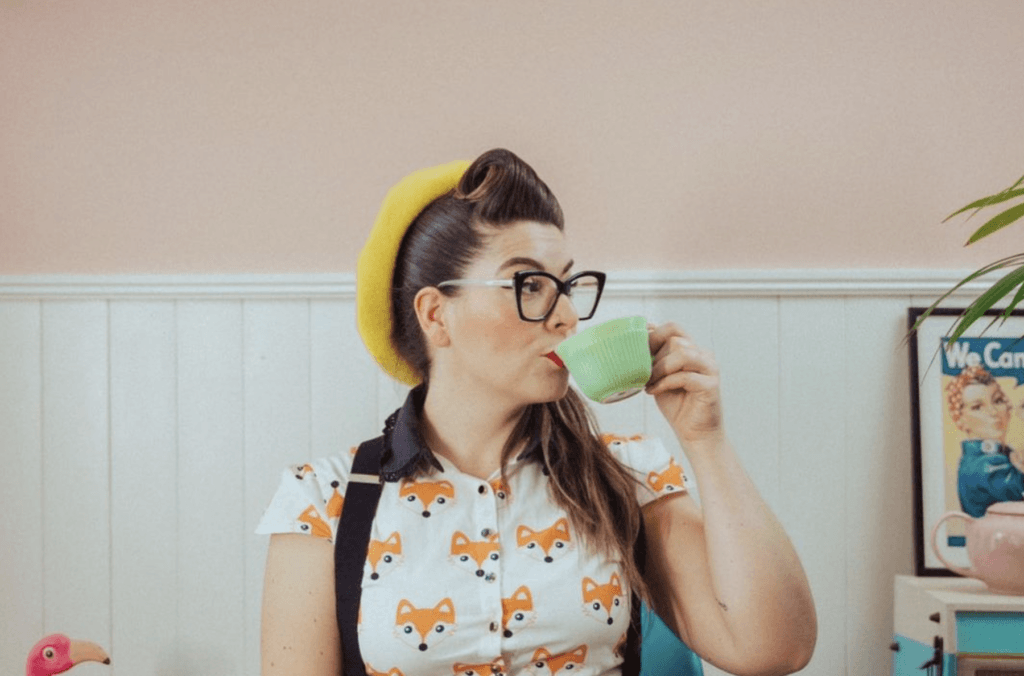 A woman in retro, 1950s clothing drinks from a green cup which she holds to her mouth