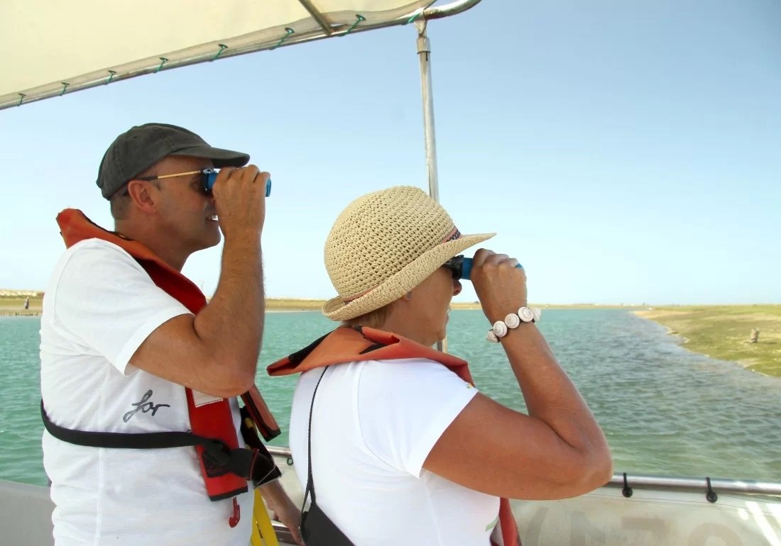 A man and woman look inland through binoculars from a boat at sea near the coast.