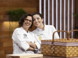 Two women, one wearing glasses, smile at teh camera as they stand, wearing chef's whites.