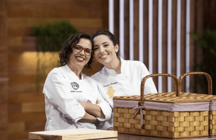 Two women, one wearing glasses, smile at teh camera as they stand, wearing chef's whites.