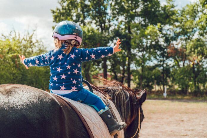 A young child, wearing a helmet, sits open armed on a horse.