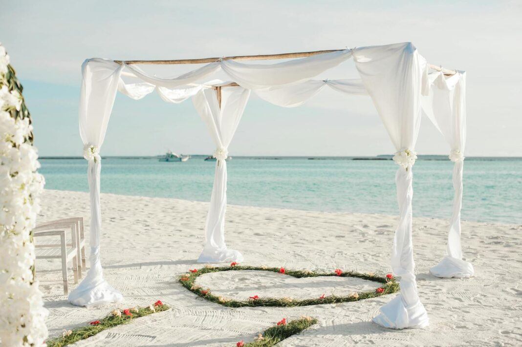 A wedding style canopy with a heart decoration on teh sand, on a beach with the sea and two boats in the background.