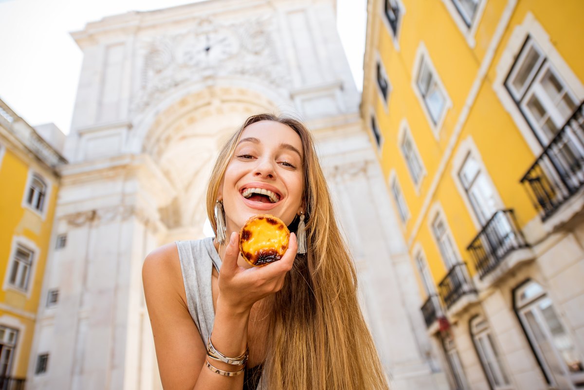 A young woman with long hair holds a pasteis da nata, as if about to eat it, with a historic archway in the background behind her.