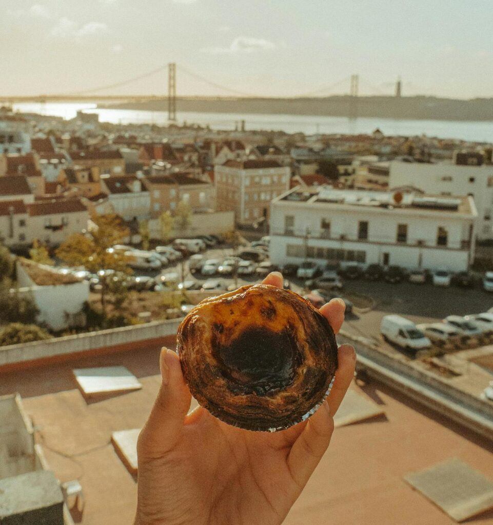 A person holds a Pastéis de Nata on a balcony with the city of Lisbon, and river visible in the background.