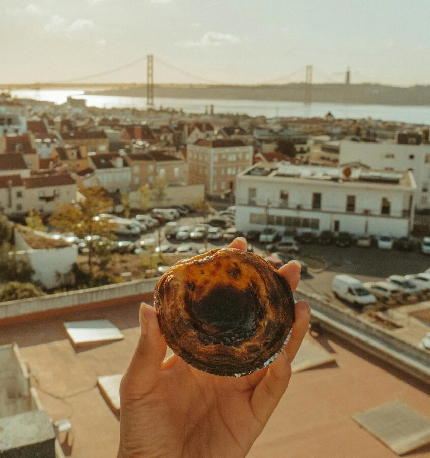 A person holds a Pastéis de Nata on a balcony with the city of Lisbon, and river visible in the background.