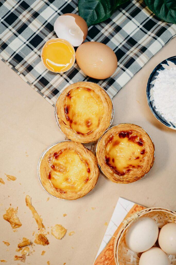 One cracked egg, and one whole seen on a counter-top with three finished, baked Pastéis de Nata.