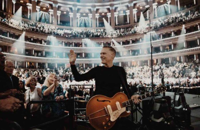 Musician Bryan Adams waves to fans with his back turned to the auditorium at a concert venue