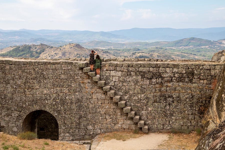 A castle wall, with steps, seen with two women standing atop teh steps looking at the countryside.