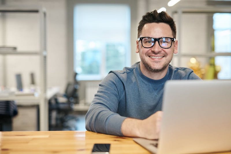 A man smiles and looks at the camera in an office as he sits at a desk with a laptop just visible