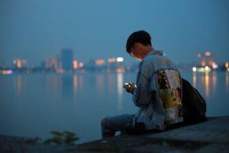 A young person hunches over a smartphone at the edge of a river with a city in teh background, in the evening.