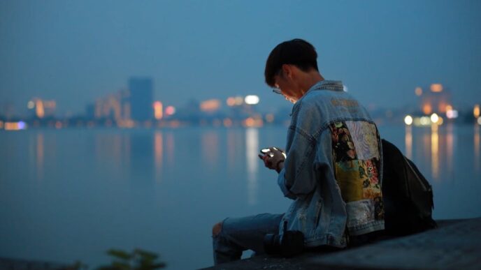 A young person hunches over a smartphone at the edge of a river with a city in teh background, in the evening.