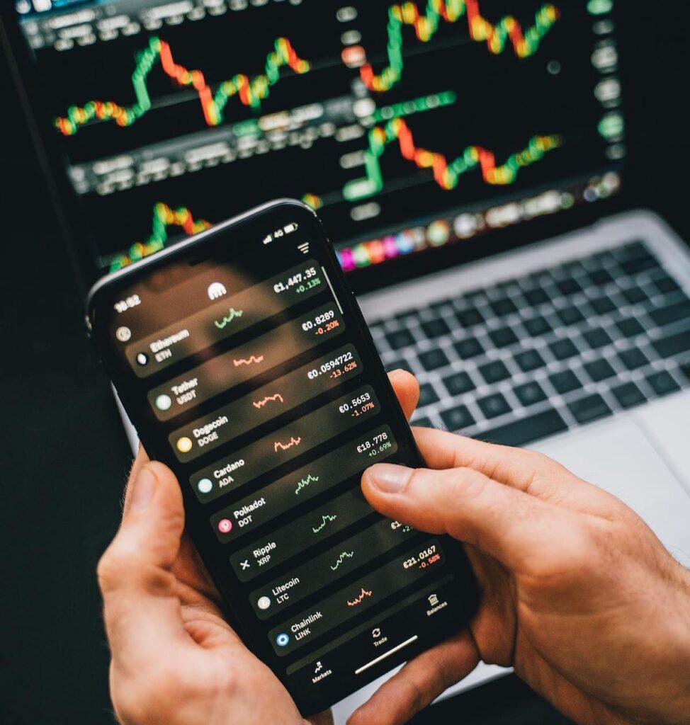 A phone with cryptocurrency prices displayed on it, with a laptop in the background, with financial charts showing on its screen.