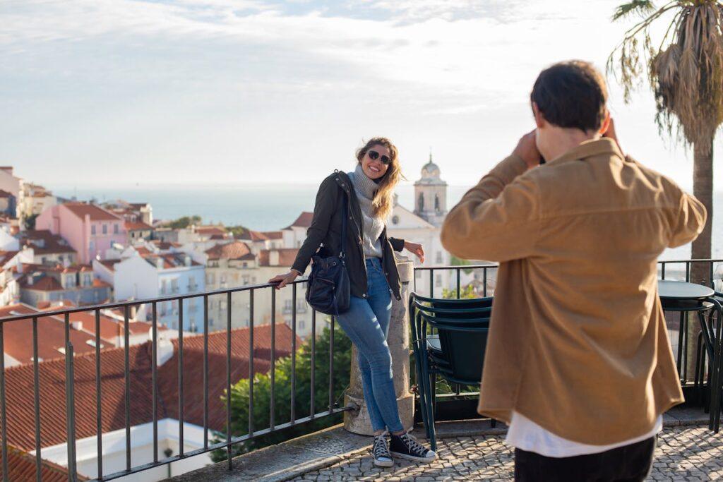 A woman poses on a balcony, outdoors, in Lisbon for a photo by a man holding a camera, with his back visible