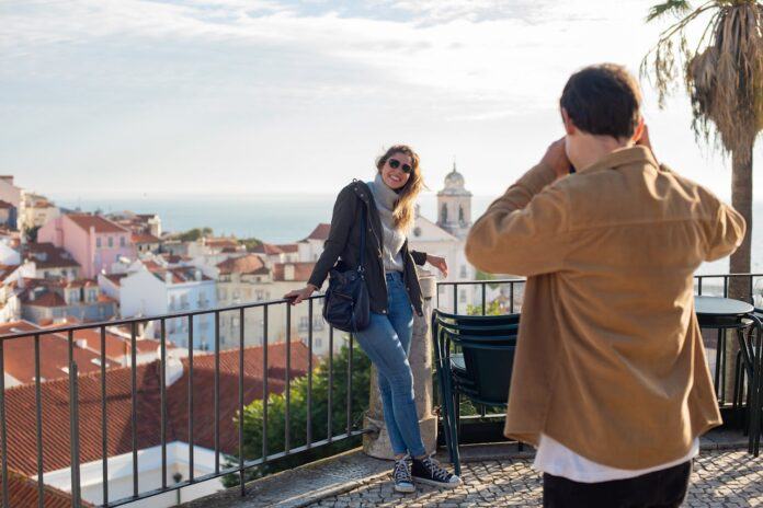 A woman poses on a balcony, outdoors, in Lisbon for a photo by a man holding a camera, with his back visible