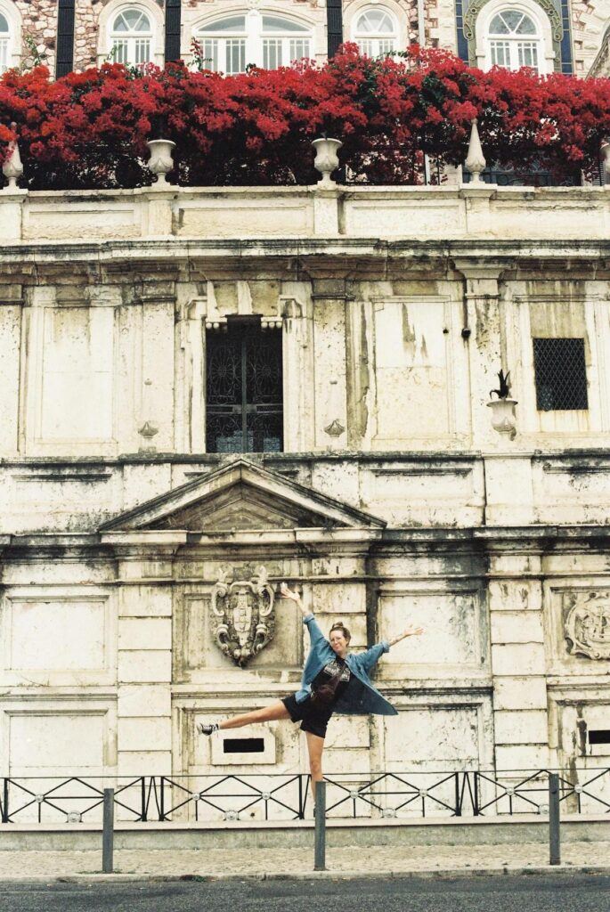 A woman dances playfully and smiles in from of teh Chafariz d’El Rei in Lisbon