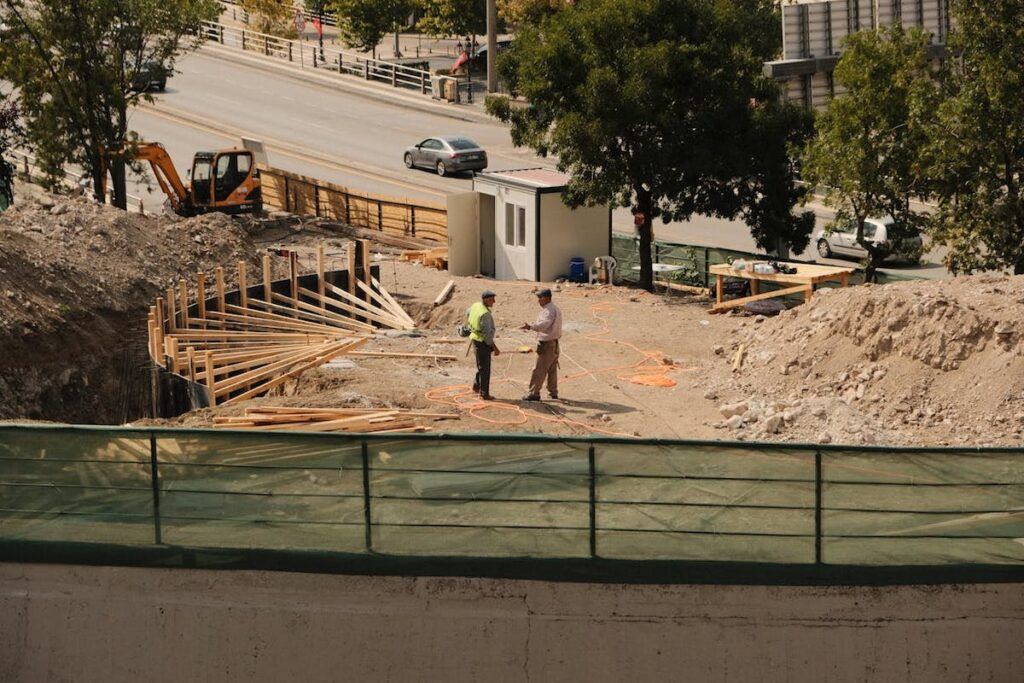 Two men talk on a building site with construction material and machinery in the photo