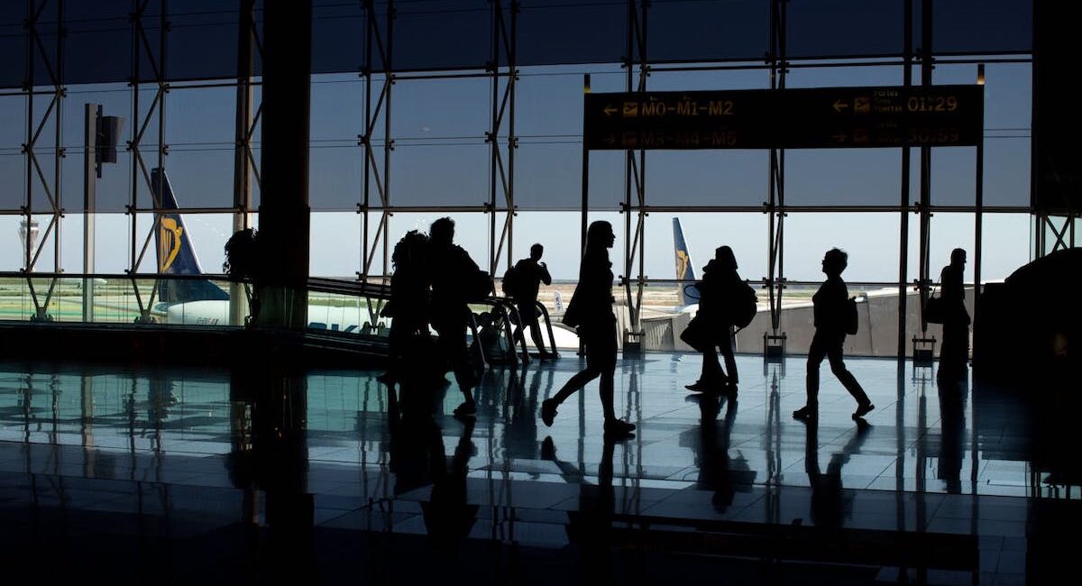 Travelers walk past a large window at an airport silhouetted against the sky with aircraft seen in the background