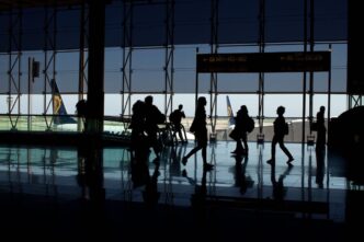 Travelers walk past a large window at an airport silhouetted against the sky with aircraft seen in the background