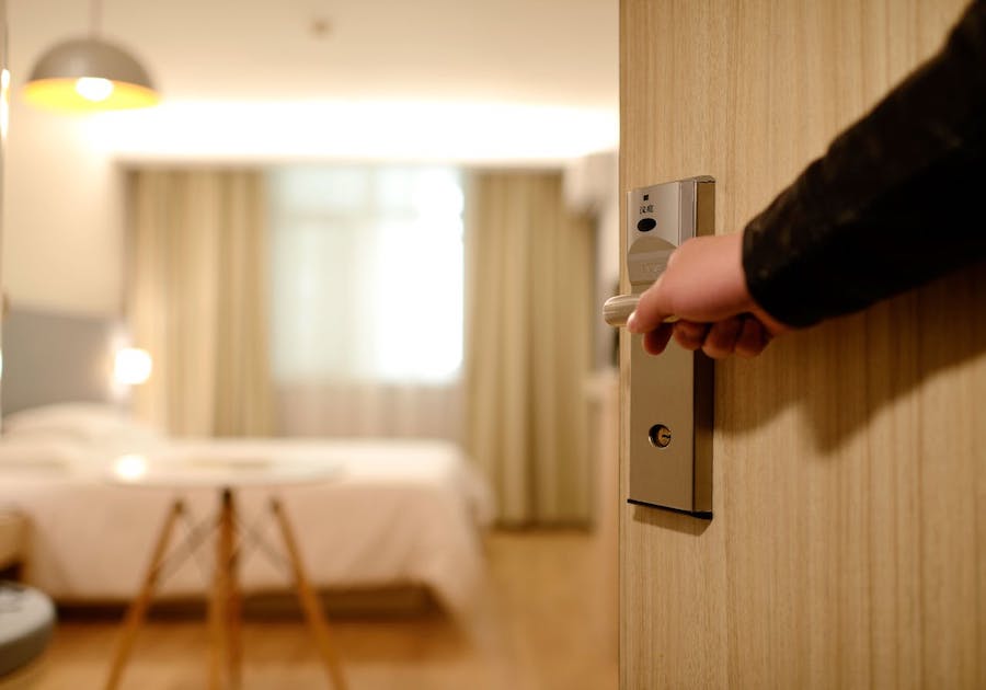 A person's hand opens a hotel room door revealing the room with a bed out of focus