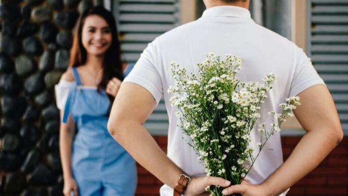 A man holds a bunch of flowers behind his back to surprise a woman waiting with a smile on her face