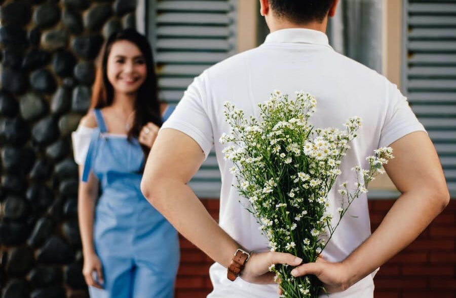 A man holds a bunch of flowers behind his back to surprise a woman waiting with a smile on her face